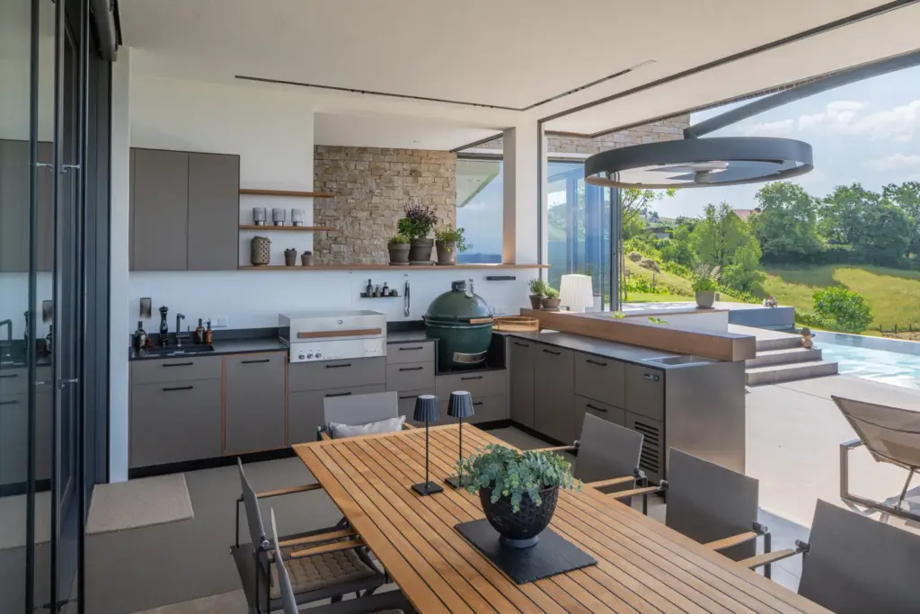 Maßgefertigte Outdoorküche mit Steinwand, Holzdetails und Blick auf Pool und Landschaft von Deutschmann