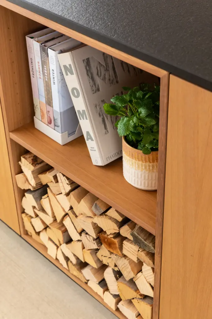 Detail einer Holzverkleidung mit schwarzer Steinplatte Kochbüchern Topfpflanze und Kaminholz im modernen Möbeldesign von Deutschmann