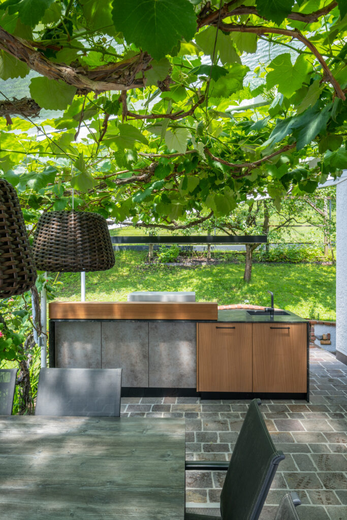 Moderne Außenküche mit grauer Front sowie zur Hälfte heller Holzfront, dunkler Naturstein Arbeitsplatte, mitten unter einer Weinrebe