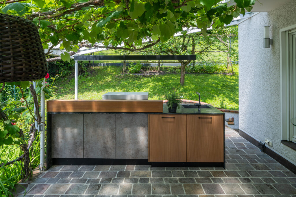 Moderne Außenküche mit grauer Front sowie zur Hälfte heller Holzfront, dunkler Naturstein Arbeitsplatte, mitten unter einer Weinrebe