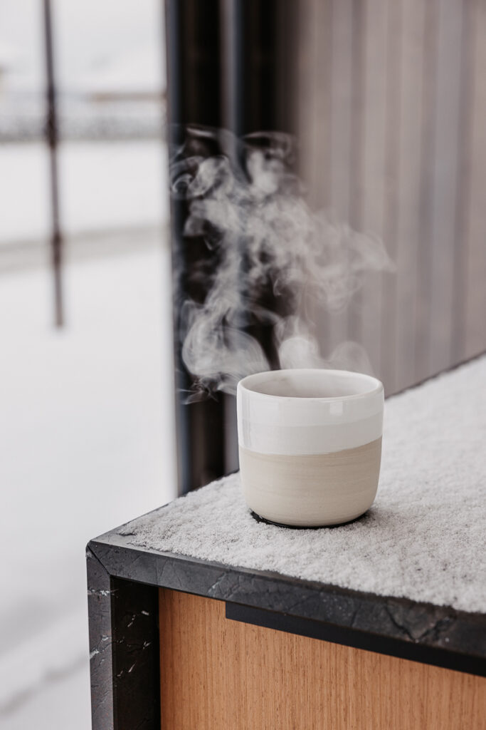 Detailansicht einer Ecke einer Outdoorküche, wo man sieht wie eine heiße, dampfende Tasse Tee auf der schneebedeckten Arbeitsfläche steht
