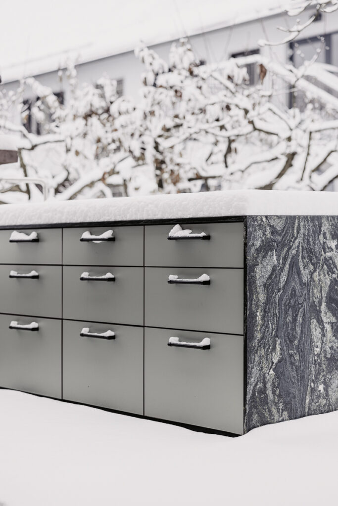 Grüne Outdoorküche mit Marmor-Arbeitsfläche welche aber mit dichtem Schnee bedeckt ist, schwarze Griffe, welche ebenso voll Schnee sind