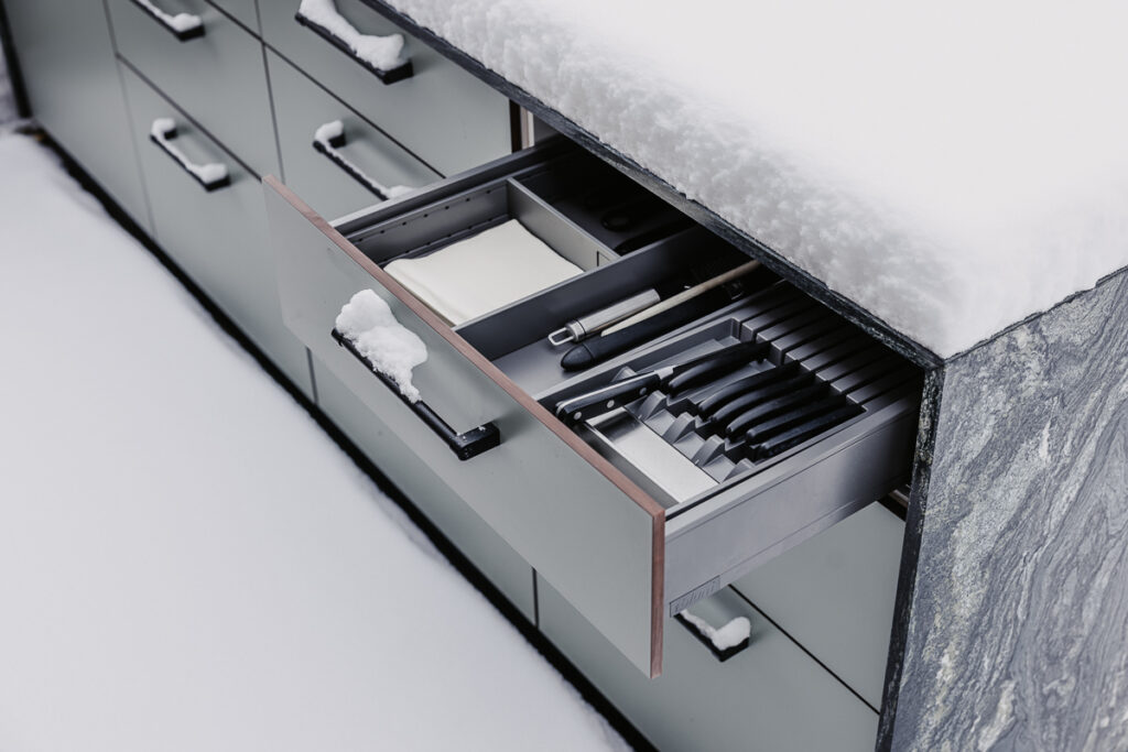 Draufansicht einer offenen Schublade einer Outdoorküche von Deutschmann. Gabeln, Messer, Servietten und weiteres Koch- & Grillbesteck darin aufbewahrt, Küche steht im Schnee und ist mit Schnee bedeckt