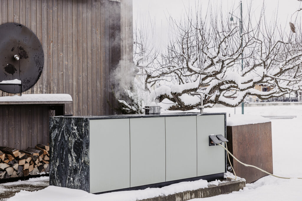 Outdoorküche mit grüner Front und Marmorarbeitsfläche, darauf steht ein dampfender Topf mit Wasser, Plug&Play Anschlüsse sind zu sehen