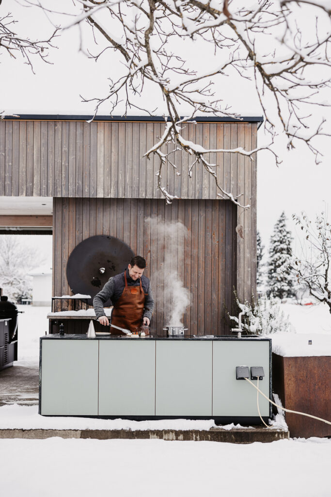 Sepp Deutschmann grillt und kocht auf der Plug & Play Outdoorküche, Gemüsespieße und Fleisch auf dem Teppanyakigrill und ein dampfender Kochtopf auf dem Indu+ Kochfeld daneben