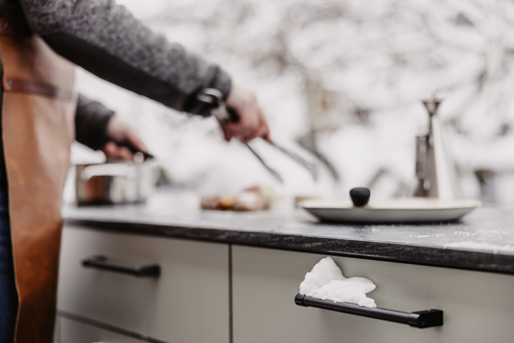Nahansicht einer Plug&Play Outdoorküche woran gerade im Hintergrund gegrillt wird. Fokus auf die schwarzen beschneiten Griffe