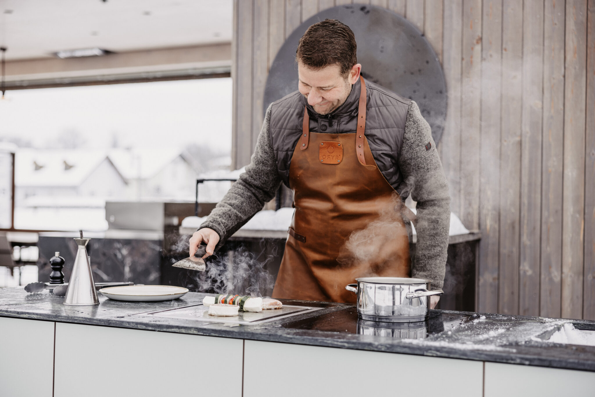 Sepp Deutschmann grillt und kocht auf der Plug & Play Outdoorküche, Gemüsespieße und Fleisch auf dem Teppanyakigrill und ein dampfender Kochtopf auf dem Indu+ Kochfeld daneben