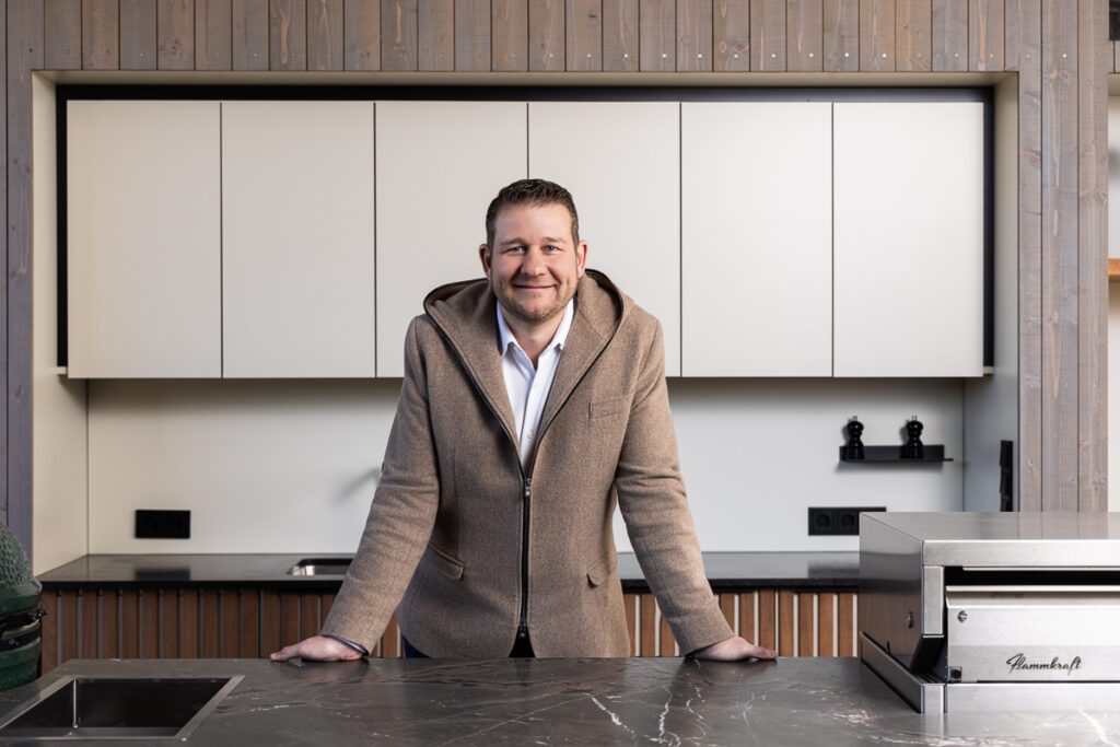 Sepp Deutschmann lehnt mit beiden Händen über eine seiner Outdoorküchen. Oberfläche dunkler, Marmorstein und Helle Fronten der Hängeschränke im Hintergrund