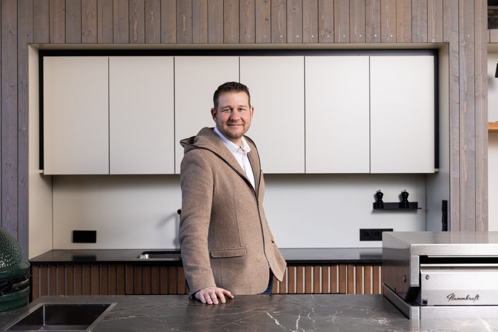 Sepp Deutschmann lehnt mit einer Hand über eine seiner Outdoorküchen. Oberfläche dunkler, Marmorstein und Helle Fronten der Hängeschränke im Hintergrund