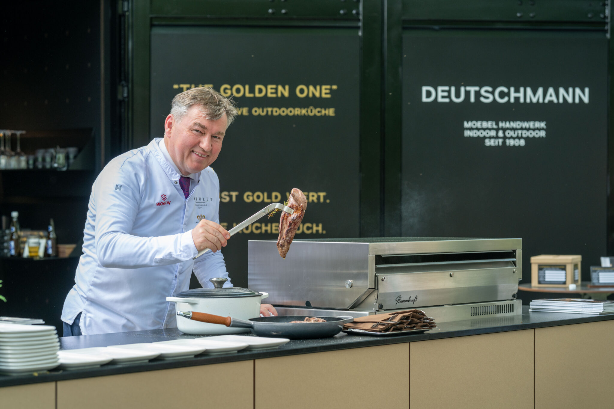 Toni Mörwald lächtelt mit einem Stück Fleisch in der Hand ins Gesicht, er steht an der Outdoorküche The Golden One von Deutschmann