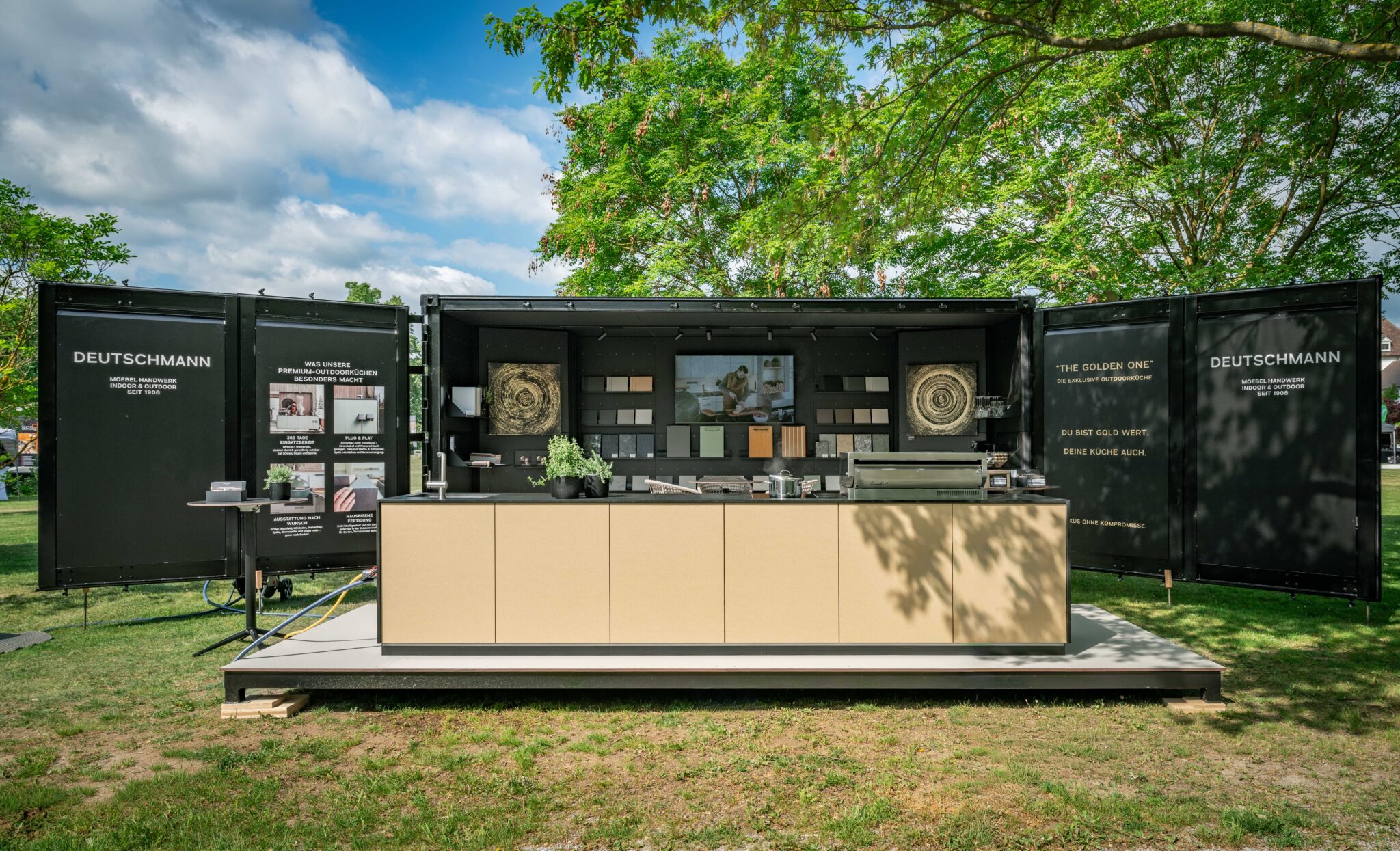 Offener schwarzer Messecontainer mit Muster darin, davor The Golden One, die goldene Outdoorküche von Deutschmann