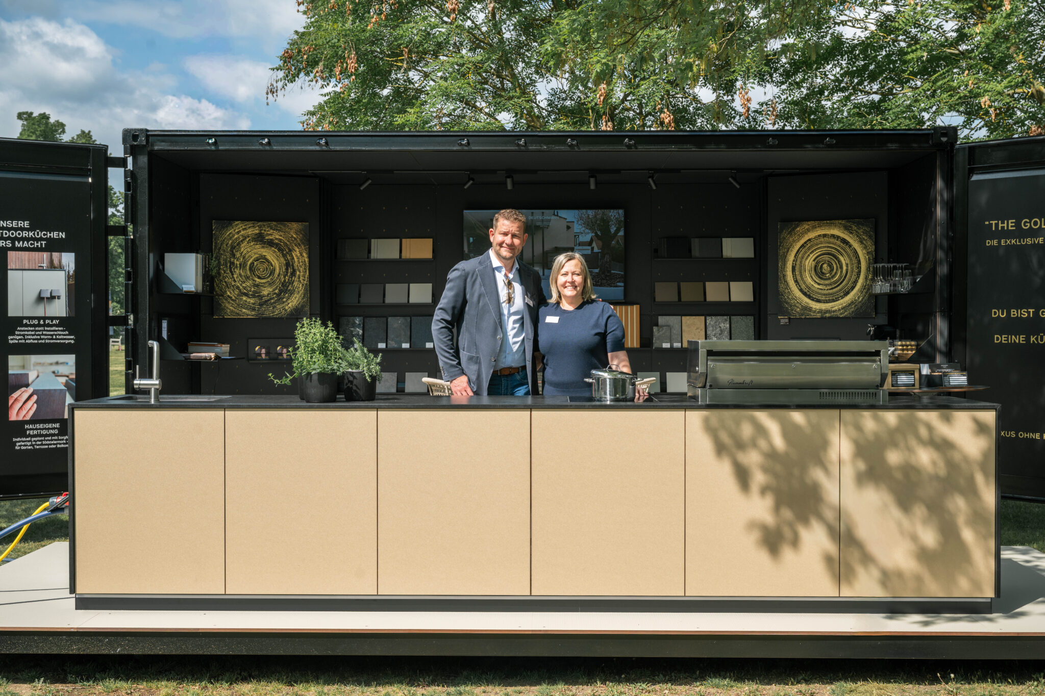Offener schwarzer Messecontainer mit Muster darin, davor The Golden One, die goldene Outdoorküche von Deutschmann, an der Küche stehend Sepp und Klaudia Deutschmann