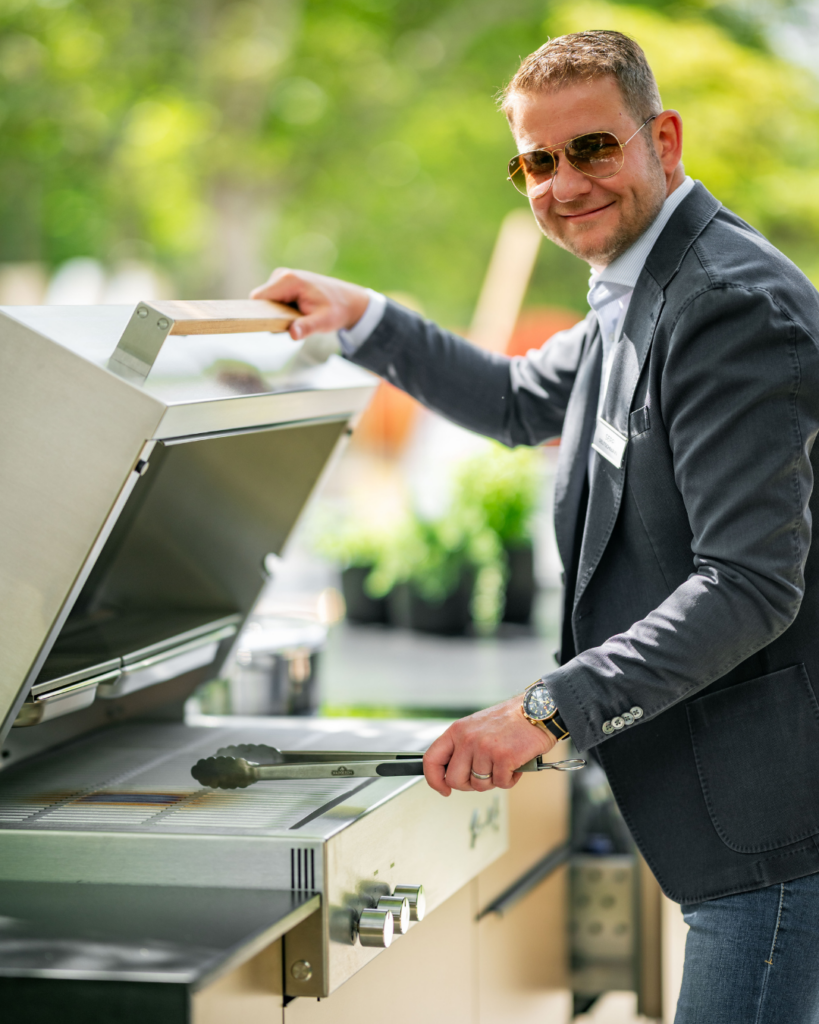 Sepp Deutschmann lächelt mit Sonnenbrille in die Kamera, stehend am offenen Flammkraft Gasgrill der Outdoorküche von Deutschmann