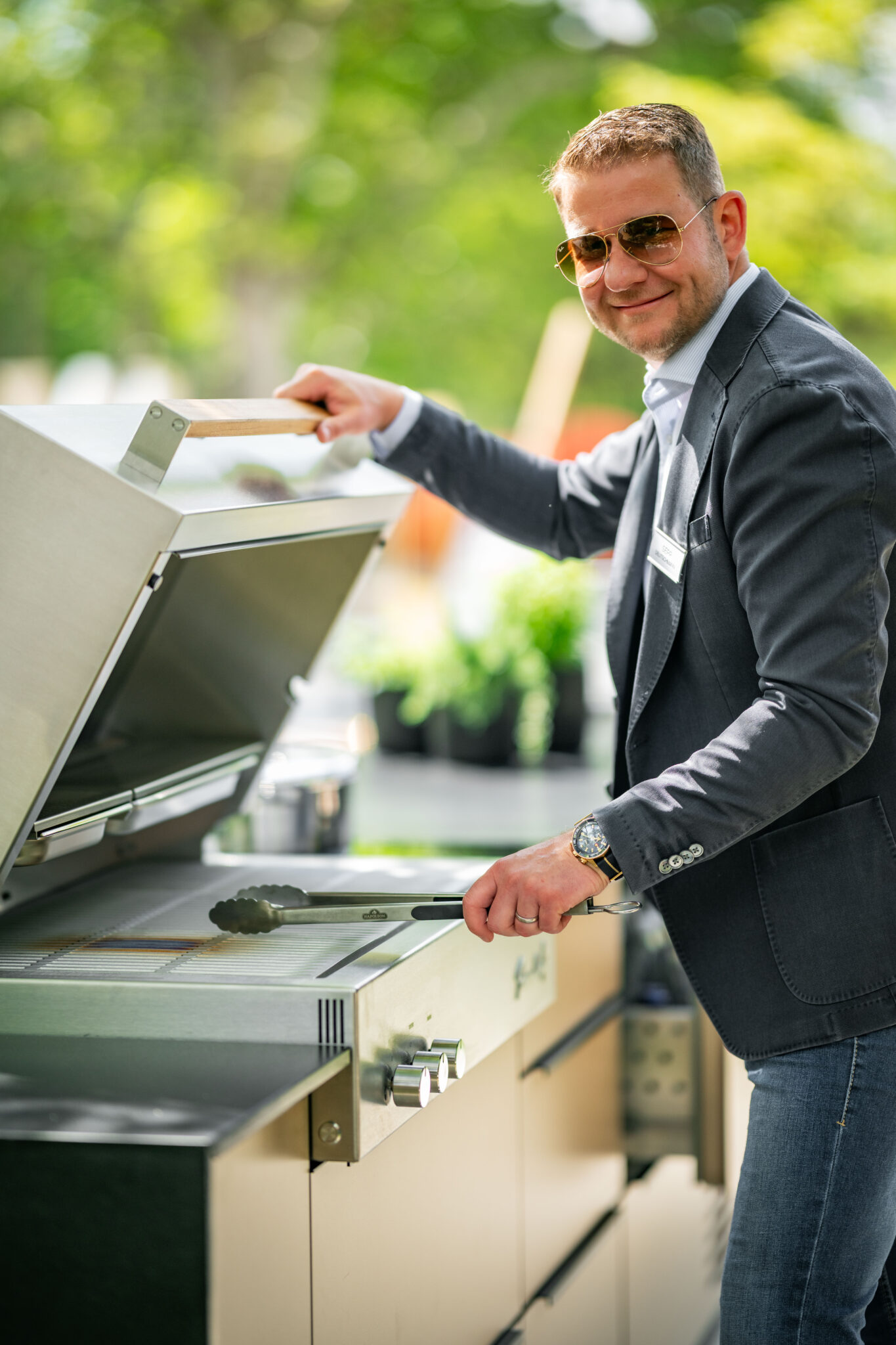 Sepp Deutschmann lächelt mit Sonnenbrille in die Kamera, stehend am offenen Flammkraft Gasgrill der Outdoorküche von Deutschmann