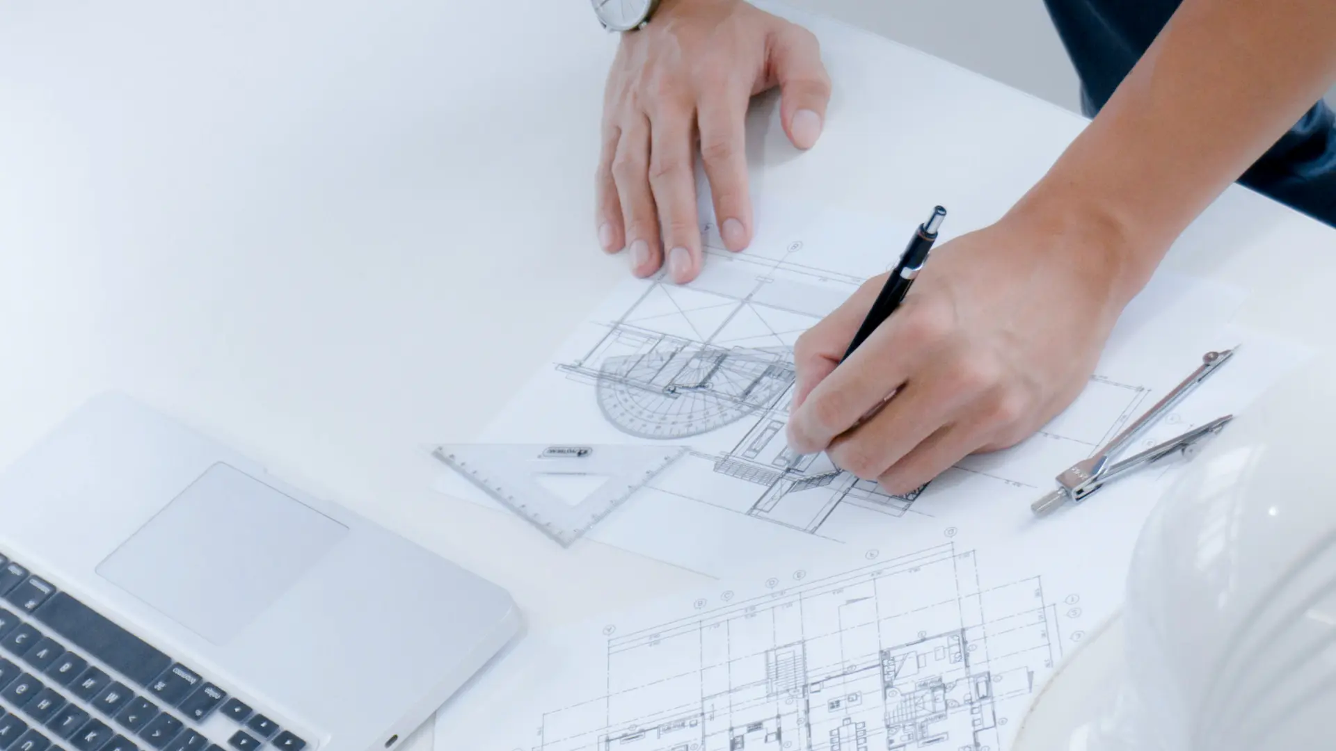 Zwei Hände auf deinem Plan, eine Hand hält einen Stift und schreibt etwas auf den Plan, daneben zwei Lineale und ein Zirkel